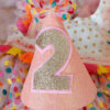 Front view of a sparkling pink 'confetti' birthday party hat, with custom golden glitter number 2 displayed on the front.